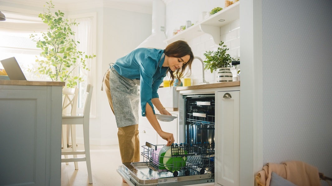 Young brunette Caucasian woman loading dirty plates into the dishwasher in a bright sunny kitchen